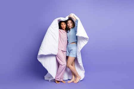 Two young women posing together playfully under a blanket, wearing stylish pajamas and standing against a bright purple backgroundの写真素材