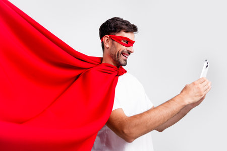 Smiling man in red superhero costume with a mask taking a selfie against a white background with expression of happiness and enthusiasmの写真素材