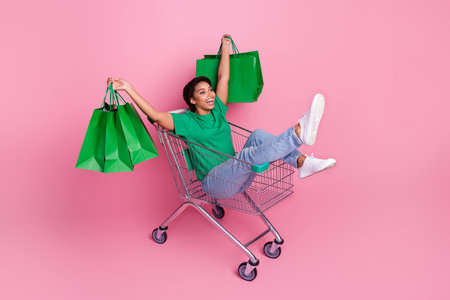 Full length photo of lovely young lady hold bags ride shopping cart have fun wear stylish green...