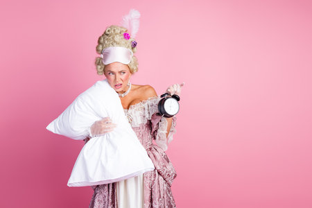 Elegant woman in historical baroque attire posing with pillow and alarm clock against pink backgroundの写真素材