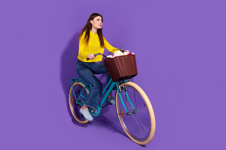 Young woman riding a stylish bicycle with a basket on a vibrant purple background, wearing a casual yellow sweater and jeansの写真素材