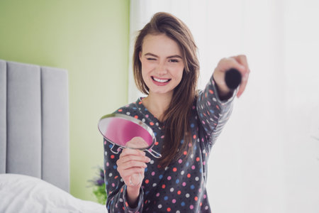 Cheerful young woman holding a mirror and a makeup brush, smiling brightly in a cozy bedroom setting, wearing polka-dotted sleepwearの写真素材