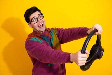 Cheerful Young Man Holding a Steering Wheel Against a Vibrant Yellow Background, Expressing...