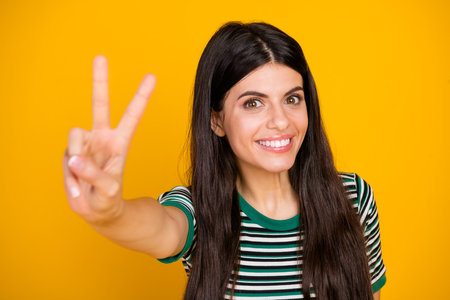 Smiling young woman showing a peace gesture against a yellow background, dressed in trendy casual...