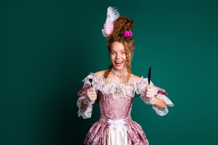Portrait of a cheerful young woman in a baroque-style pink dress holding cutlery against a green...