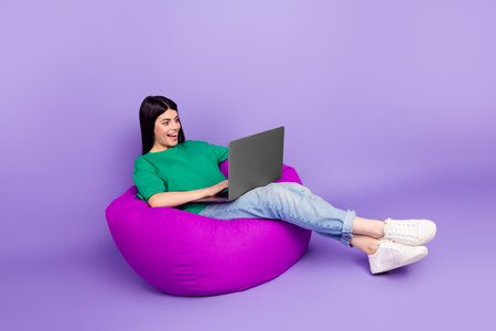 Cheerful young woman enjoying leisure time on a vibrant bean bag with a laptop in casual setting on a vivid violet background.の写真素材