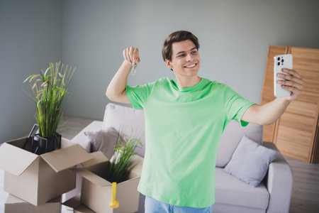 Young man taking a selfie while holding keys, celebrating new apartment in modern home interior with unpacked boxes and houseplants.の写真素材