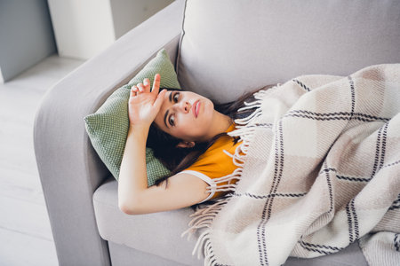 Young woman relaxing on cozy sofa in stylish t-shirt covered with plaid blanketの写真素材