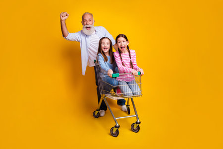 Excited grandfather pushes two happy young girls in a shopping cart against an energetic yellow backgroundの写真素材