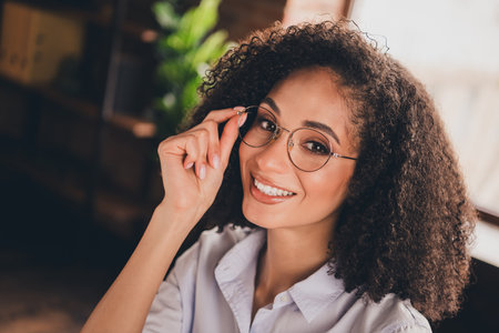 Confident young businesswoman with curly hair and glasses smiling in a modern office settingの写真素材