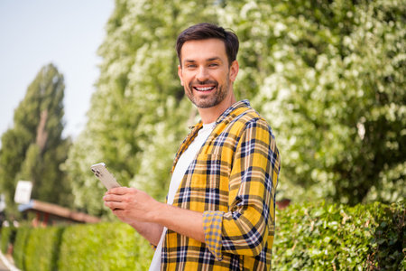 Young trendy man in checkered shirt smiling and holding smartphone while enjoying a sunny day outdoors in a scenic park.の写真素材