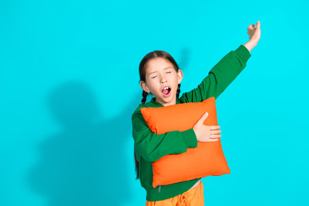 Young girl yawning while holding an orange pillow against a turquoise background, wearing a green hoodieの写真素材
