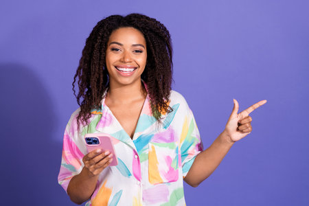 Happy young woman with curly hair smiles confidently, holding a smartphone and pointing with a hand...