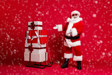 Full length photo of retired old man white beard recommending sled many giftbox best choice wear santa x-mas costume coat sunglass headwear black boots isolated red color backgroundの写真素材
