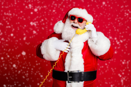 Photo of pensioner old man grey beard hold wired telephone talk snow maiden receive compliment hand chest wear x-mas santa costume gloves coat sunglass cap isolated red color backgroundの写真素材