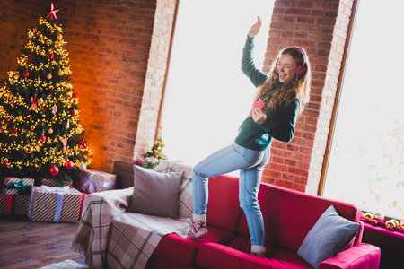 Full length photo of teen girl dance have fun on sofa dressed green christmas garment tradition atmosphere decor vibe house indoors roomの写真素材