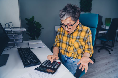 Photo of nice retired woman confused nervous use calculator wear plaid shirt comfortable modern...