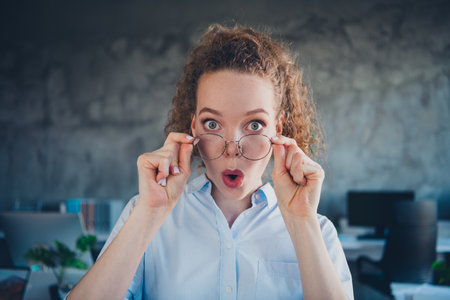 Surprised young woman in formalwear at office adjusting glasses, expressing amazement and curiosity...