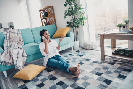 Full size photo of pretty young girl sit carpet floor speak phone wear shirt modern interior apartment indoorsの写真素材