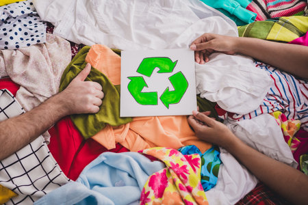 Hands holding recycling sign amidst colorful clothes, promoting eco-friendly fashion and sustainable living with a diverse touchの写真素材