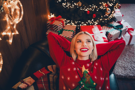 Photo of young blonde woman dreamy look relax hands behind head dressed christmas sweater day light home party decoration interior roomの写真素材