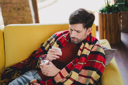 Handsome young man wrapped in blanket holding medication while relaxing at home on a cozy autumn day, enjoying the natural lightの写真素材