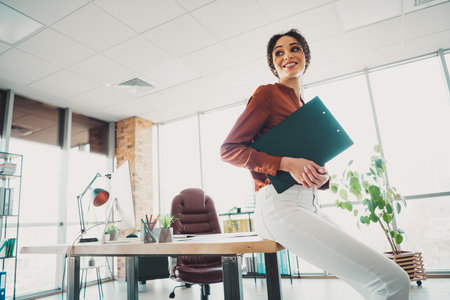 Young professional businesswoman in casual office attire holding documents while posing in a modern workplace with natural daylightの写真素材