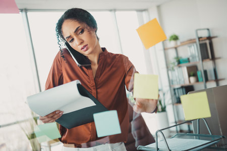 Young professional woman multitasking in a busy office environment, interacting with notepads and sticky notes under natural daylightの写真素材