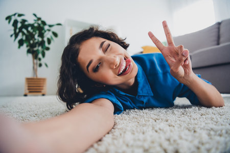 Young woman in casual shirt smiling and making a peace sign gesture while relaxing on a white carpet in a cozy living room.の写真素材
