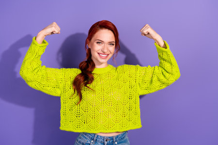 Confident young red-haired woman showing strength gesture against purple background