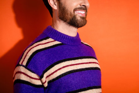 Smiling young man wearing striped sweater against vibrant orange background, showcasing bearded...