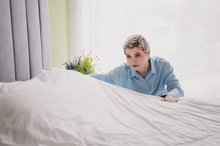 Young woman making her bed in a bright sunlit bedroom, reflecting a moment of calm and routine in a cozy indoor home environmentの写真素材