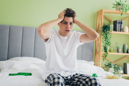 Young man looking stressed in casual pajamas sitting on a comfy bed in a well-lit cozy bedroom at homeの写真素材