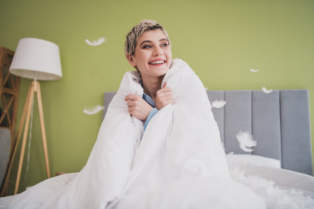 Young woman smiling in bed wrapped in a soft white duvet enjoying a cozy morning at home in a bright and stylish bedroom.の写真素材