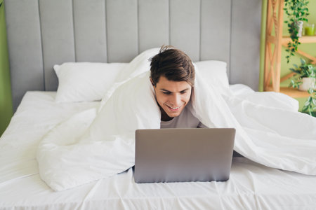 Young man under a duvet using a laptop with a cheerful expression in a cozy bright bedroom
