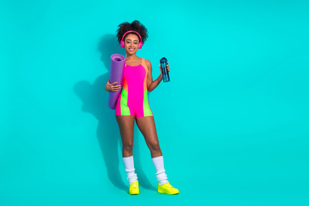 Sporty Young Woman in Stylish Jumpsuit with Colorful Design Holding Yoga Mat and Water Bottle in...