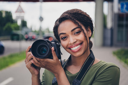 Charming young photographer with afro hairstyle smiling in urban setting holding camera outdoors on a sunny dayの写真素材