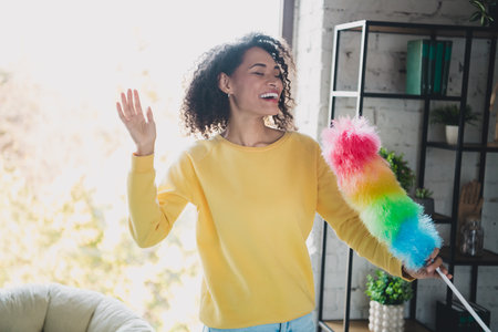 Smiling woman in yellow sweatshirt dusting her home with colorful duster and enjoying a joyful...