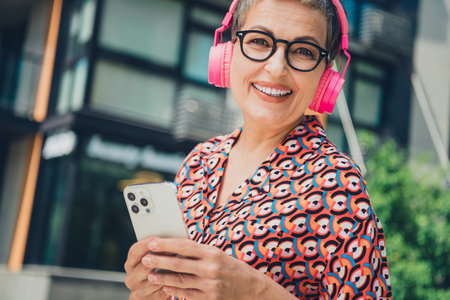 Elderly woman enjoying music with pink headphones and smartphone outdoors during a sunny day in the...