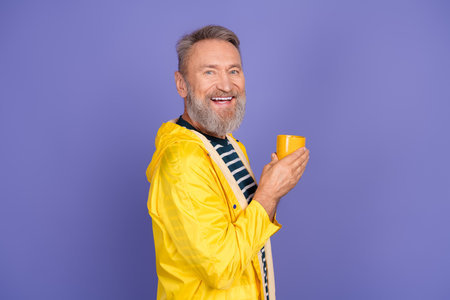 Smiling mature man in a yellow raincoat holding a cup against a purple background, showcasing...