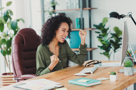 Young joyful businesswoman celebrating success at her work desk in a bright and modern office with natural daylightの写真素材
