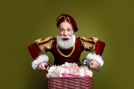 Elderly nobleman in renaissance attire rides bicycle with flowers in basket on khaki background, blending vintage fashionの写真素材
