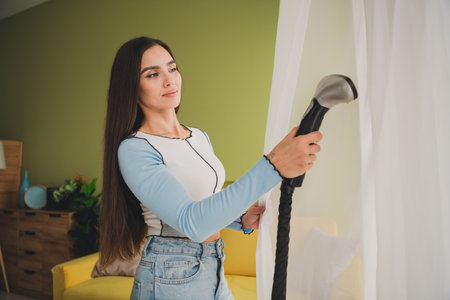 Young brunette woman using a steamer to tend to curtains in a cozy living room setting, reflecting modern lifestyle and domestic careの写真素材