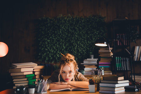 Young woman studying indoors at home with books late in the evening, wearing glasses and checkered shirt, surrounded by booksの写真素材