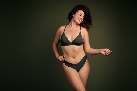 Confident young woman in stylish dark lingerie posing gracefully against a khaki background, embracing natural beautyの写真素材