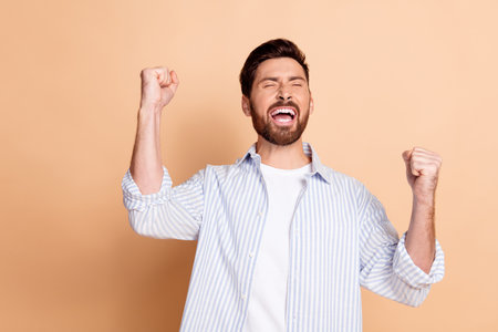 Excited young man celebrates success with a joyful expression and raised fists on a neutral beige background representing positivityの写真素材