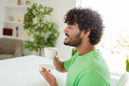 Young man enjoying beverage indoors with delighted smiling expression surrounded by cozy home decor and natural daylightの写真素材