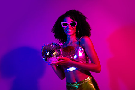 Portrait of a vibrant young woman in metallic attire holding a disco ball against neon lights, evoking night club energy and retro partyの写真素材