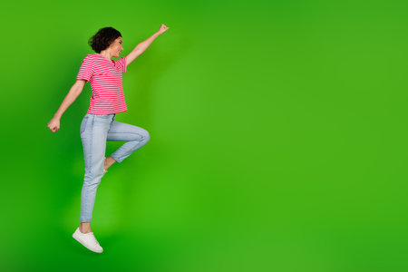 Young energetic woman jumping joyfully in green background wearing casual striped outfit and showcasing a lively attitudeの写真素材