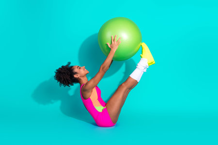 Young athletic woman balancing green exercise ball on her feet, wearing vibrant sportswear, expressing vitality and a healthy, active lifestyle against a turquoise background.の写真素材